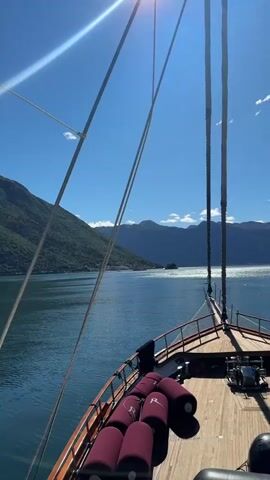 The video captures a view from the bow of a sailing ship on a sunny day. The deck is wooden and has a dark wood railing. Seven dark red cylindrical flotation devices with the letter "R" are placed along the railing. The ship's rigging, consisting of thick ropes and the mast, extends upwards towards the bright sun in a clear blue sky. A body of water stretches out to the horizon, surrounded by mountains covered in green vegetation. Another mountain range is visible in the distance. The water is slightly rippled and reflects sunlight. A small island is visible in the middle of the water.