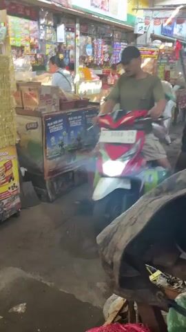 A man wearing a dark baseball cap and a green t-shirt rides a red and white motorcycle through a crowded marketplace. He is wearing khaki shorts and appears to be carrying goods on the back of the motorcycle. The motorcycle's headlights are on, illuminating the path ahead. The marketplace is filled with stalls and shelves stocked with various items, and other people can be seen in the background. The overall atmosphere suggests a busy and active commercial environment.