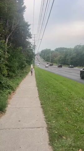 A person is walking on a sidewalk next to a busy road. The person is walking away from the camera and is on the left side of the frame. The road is on the right side of the frame and has cars driving on it. The sky is overcast and there are trees on the left side of the frame. There are also power lines running above the road.