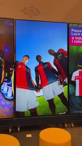 The video shows three large vertical display screens featuring promotional images. The central screen showcases three male soccer players in the red, white, and blue uniforms of Genoa. They are standing on a soccer field under a bright blue sky. To the left, the display screen shows a stylized neon display. To the right, the display screen shows an advertisement for Pulsee Luce e Gas, featuring the Genoa soccer team uniforms again. The video then focuses on the center screen, now showing a woman in a Genoa tracksuit walking down a street in the city of Genoa. Behind her is a man, also in the same tracksuit, walking away. The room has two bright yellow round stools in the foreground.