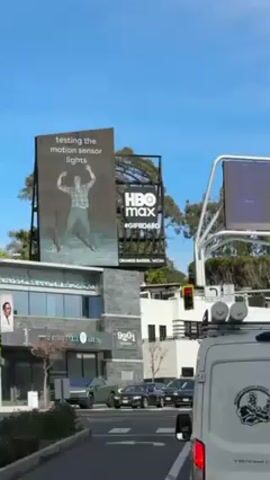 From a vehicle on a road at an intersection, a digital billboard is seen to the left. The billboard promotes HBO Max, with the text “testing the motion sensor lights” and an image of a man, with his arms raised. There are trees, buildings, cars, and traffic signals nearby. The sky is blue. There is another digital billboard to the right of the first one. A white work van is in the foreground.