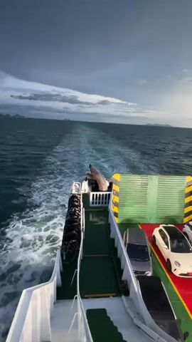 The video shows a view from the deck of a ferry sailing on a body of water. The ferry is carrying several cars on its deck. The water behind the ferry is churned into a white wake, indicating the vessel is moving forward. In the distance, landmasses, likely islands, are visible on the horizon. The sky is cloudy with dark, heavy clouds suggesting an approaching storm or the end of the day. The lighting indicates either early morning or late evening. The overall mood is somewhat dramatic due to the weather conditions. There is no visible text in the media.