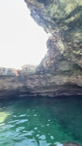 A wide shot captures a person in a black bikini and a tan hat sitting in a small white boat. The boat is in clear, turquoise water next to a rocky cliff face. The water is illuminated by sunlight, and the rocks are textured and varied in color. The sky above is a clear, bright blue. Another person, with long blonde hair, sits in the boat to the left, also in a bikini. The camera pans slightly to the right, showing more of the rocky overhang and the water below. The overall scene is one of a sunny day spent on the water near a coastal cave or rock formation.