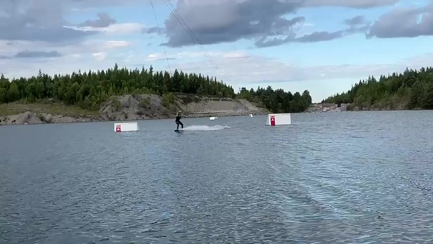 A person wakeboards on a lake. They are wearing dark clothing and a helmet. The wakeboarder is being pulled by a cable that is attached to a tower. There are obstacles in the water, which appear to be ramps or jumps. The lake is surrounded by trees and hills. The sky is cloudy.