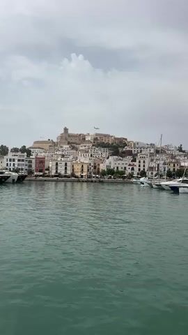 The video shows a wide shot of a coastal town built on a hillside. The town is characterized by whitewashed buildings with some colorful accents, clustered together. At the very top of the hill, there is a prominent fortified structure, possibly a castle or a cathedral. In the foreground, the turquoise water of a harbor is visible, with several yachts and sailboats docked. The sky is overcast with some clouds. An airplane is seen flying in the sky above the town. The camera appears to be moving slightly, creating a gentle pan across the scene. The overall atmosphere is serene and picturesque, capturing a typical view of a Mediterranean coastal settlement.