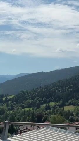 The video shows a panoramic view of a mountainous landscape with a dense forest covering the slopes. A few scattered houses are visible in the lower part of the frame, nestled within the greenery. The sky is blue with scattered white clouds. A metal railing is in the foreground, suggesting the video was filmed from a balcony or elevated viewpoint. The camera pans slowly to the right, revealing more of the forest and the undulating terrain. The overall impression is one of a serene and natural environment.