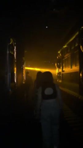 The video shows a group of people walking down a dimly lit corridor. The corridor appears to be flanked by theatrical set pieces on either side, possibly representing train carriages or ornate buildings. There are strong beams of orange and yellow light casting shadows and creating a foggy atmosphere. The people are walking away from the camera, their faces mostly obscured by shadow. The overall mood is mysterious and cinematic.