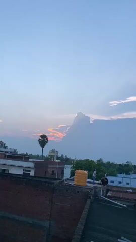A vertical, low-angle video shows rooftops of buildings under a hazy, pale blue sky with wispy clouds. A single tall, slender palm tree stands to the left of center. In the distance, a water tower is visible. The sun appears to be setting behind a large, dark cloud on the right, casting a warm orange glow on some smaller clouds in the sky. Two people are standing on a rooftop in the mid-ground, and a yellow water tank is also present. The foreground consists of a red brick roof and the side of a brick building. The video is a time-lapse, with subtle changes in the clouds and light.