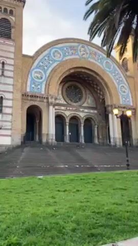 The video shows a low-angle, panning shot of a church from a park across the street. The church is a large building with a tan brick facade, a large arched entryway with mosaic tile decorations, and a tall bell tower to the right.  A wide set of steps leads up to the church entrance. In front of the church is a grassy area with scattered litter. Palm trees and lamp posts are situated around the green space. Across the street from the church, buildings can be seen, along with trees, lamp posts, and painted artwork. The sky is overcast with some glimpses of blue. No people are visible in the shot.