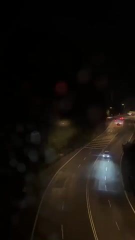 The video shows a dark highway at night with cars driving on it. There are some trees on either side of the road and streetlights illuminating the scene. The camera seems to be positioned at an elevated viewpoint, possibly from a bridge or higher building. A voice can be heard in the background. The video seems to be recorded in poor weather conditions, as there are visible water droplets on the lens.