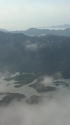 The video shows an aerial view of a mountainous landscape with several islands dotting a body of water. Clouds drift across the scene, obscuring parts of the mountains and islands. The overall lighting suggests it is daytime, possibly early morning or late afternoon, with the sun filtering through a hazy sky. The perspective is from an aircraft, as indicated by the presence of an airplane engine in the lower right portion of the frame in some shots. The terrain appears lush with green vegetation, and the water is a muted gray-green color. The distant horizon shows more mountains and hints of a coastline or city. The video captures the dynamic nature of the landscape as clouds move and the light changes.