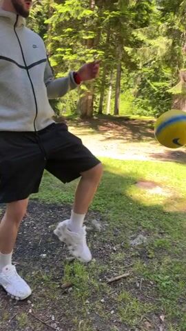A person is in a forest, throwing a yellow and blue ball in the air. They are wearing black shorts, a white hoodie, white socks, and white sneakers. The person is captured mid-jump as they throw the ball. The environment is full of tall green trees.