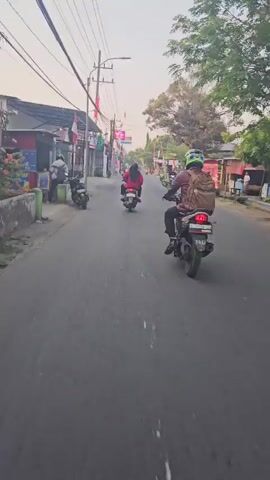 The video captures a motorcycle perspective moving along a paved road lined with buildings and trees. Several motorcycles are visible on the road, with some individuals riding them. On the left side of the road, there are buildings, some with awnings and signs. Red and white flags are displayed along the street, suggesting a festive or national occasion. A red food cart is parked on the right side of the road. The sky is bright, indicating daytime. The overall scene depicts a street in a populated area, possibly a town or village, with moderate traffic and commercial activity.