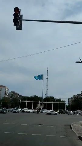 A traffic light is visible at the top left of the frame. In the center of the frame, a blue flag with a yellow sun and eagle is waving on a flagpole. Behind the flag is a tall transmission tower. In the foreground, cars are driving through an intersection. The sky is cloudy.