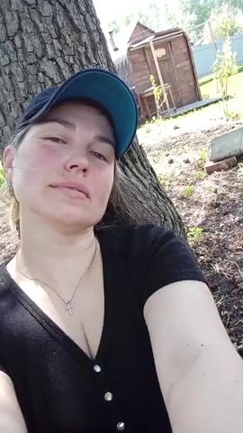 A woman is sitting with her back to a tree, looking up into the camera. She is wearing a black v-neck shirt, a silver necklace with a cross pendant, and a blue and black baseball cap. She smiles. Behind the woman is a small wooden outhouse, a grassy yard, and trees. The sun is shining brightly.