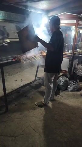 The video shows a person standing and holding a square object, possibly a fan, over a grill. The grill is filled with skewers of food, and smoke is rising from it. The scene appears to be outdoors at night, with a street in the background and a food stall to the right. Another person is partially visible in the background. The ground is concrete, and there are several trash bags on the right side of the frame. The overall impression is of a street food vendor preparing food.
