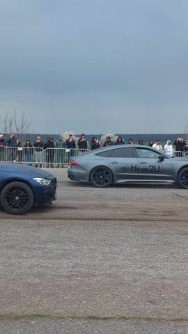 The video shows a blue BMW sedan doing a burnout on an asphalt surface, creating smoke. The car is accelerating away from the camera. A crowd of people is standing behind a barrier, watching. There is a green field in the background, and the sky is overcast. Another vehicle, likely another car, can be seen further down the road also accelerating away.