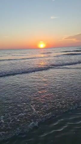 The video shows a sunset over a beach. The sun is low on the horizon, casting an orange glow across the sky. Gentle waves are rolling onto the sandy shore, creating a peaceful and serene atmosphere. The sky is a gradient of blue and pink, with a few wispy clouds. The water is a deep blue, reflecting the colors of the sunset. The camera is positioned at ground level, capturing the scene from the perspective of someone standing on the beach. The video is shot in portrait mode.
