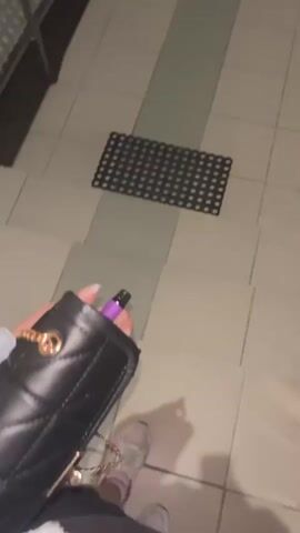 A first-person perspective video shows a person descending a tiled ramp at night, holding a black handbag and a purple vape pen. A black doormat is on the ramp. The ramp leads to a paved area with a crosswalk and street, illuminated by streetlights. Buildings are visible in the distance, including one with two towers. Cars are seen driving on the street.
