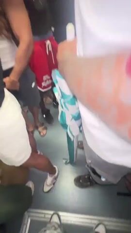 The video captures a close-up view of a person speaking directly into the camera, with their mouth open and teeth visible. The person is wearing a white shirt with red accents, and a red bandana with a white pattern is visible around their neck. In the background, other people are present, and the environment appears to be dimly lit, possibly an indoor gathering. The camera is held at a low angle, and the image is somewhat blurry, suggesting movement. The person is speaking, but their words are not transcribed.