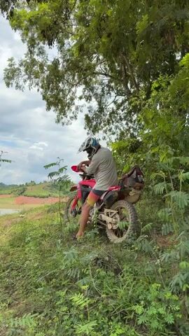 A person wearing a helmet and casual clothing sits on a dirt bike, which is positioned on a grassy incline near a body of water. The person is facing away from the camera, leaning over the handlebars. The dirt bike is red and white with some mud on its wheels. Trees and foliage surround the area, with a cloudy sky overhead. In the distance, rolling hills are visible. The person then starts the motorcycle and rides it up the incline, disappearing from view.