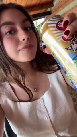 A young woman in a white sleeveless shirt with buttons is drinking from a blue and gold can of cerveza. She holds the can with her right hand, showing dark red nail polish and multiple rings. The camera is close to her face, focusing on her as she drinks. She has light brown hair and a gold necklace with the letter "R." The background includes a wooden ceiling, a brick wall, and some decorative items. The lighting is bright.