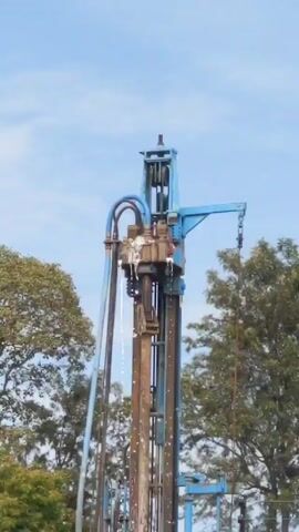 A static, medium shot shows a blue well-drilling machine framed by trees and a light-blue sky. Water is visibly spraying from the machine, which is tall and dominates the frame. There are no people visible. The trees provide a natural backdrop to the industrial machinery.