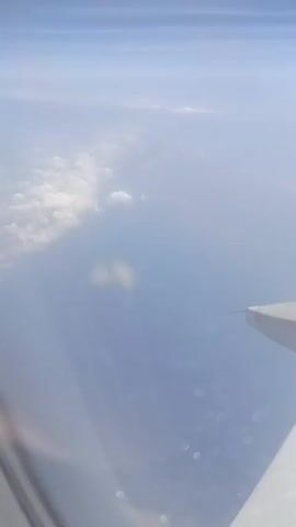 The video shows an aerial view from an airplane window. The left side of the frame shows a hazy blue sky with scattered white clouds. The right side of the frame features a portion of the airplane's wing, including several flaps and the wingtip. The wing is white with visible rivets and markings. The perspective suggests the plane is in flight, moving through the sky.