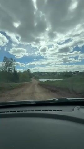 The view from inside a car traveling on a bumpy dirt road in a rural area. On either side of the road is green grass and trees. A small pond can be seen to the right of the road, and a small village can be seen in the distance. The sky is blue with white clouds. The car is moving forward, and the view is shaky due to the bumpy road.