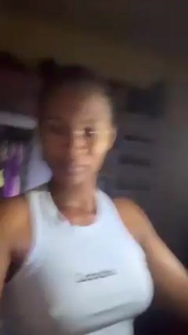 A woman with dark skin, wearing a light blue tank top with the word "Labubu" on it, is looking directly into the camera. She appears to be indoors, possibly in a room with a dark background. The lighting is dim, but a brighter light source is slightly behind her. The woman seems sweaty. The camera angle suggests she is holding the camera at arm's length, likely taking a selfie or short video.  A few clothing items are vaguely visible in the background.