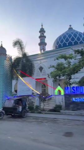 The video pans from left to right, showcasing a mosque and its surroundings. In the foreground, the sign for "MASJID AGUNG BAITURRAHIM LHOKSUKON ACEH" is prominently displayed, illuminated by blue neon lights. The mosque itself is visible, featuring a large blue dome and two minarets, also lit with blue lights. Trees with lush green foliage partially obscure the mosque's facade. In the background, a white car is parked on the left, and a street runs horizontally across the scene. Further to the right, a yellow truck is visible, along with a small market stall displaying fruits and vegetables. A motorcycle is parked near the market. The sky is overcast, suggesting it is either early morning or late evening. The overall impression is of a public space in a town or city, with religious and commercial activity coexisting.