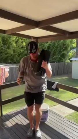 A man in a baseball cap, gray t-shirt, black shorts, and gray Crocs dances on a wooden deck holding a black JBL speaker and a can, possibly a drink. He stands on a wooden deck with a covered pergola structure, green lawn, a white bucket, and a clothesline visible. The man moves rhythmically to the music emanating from the speaker.