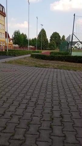 This is a video of an outdoor recreational area. The foreground is a paved walkway made of interlocking gray bricks with some grass growing between them. Beyond the walkway is a manicured lawn with a low hedge. Further back, a sports field is visible, enclosed by a tall net fence. There are also several trees and some buildings in the background, along with tall light poles. The sky is partly cloudy with a bright, blue expanse, suggesting it is daytime. The overall impression is a well-maintained public space, likely a park or school grounds.

The video appears to be taken from a low angle, possibly from someone walking or pushing a stroller, as indicated by the slight motion of the camera. There are no people visible in the frame. The scene is peaceful and calm. The time of day appears to be late afternoon or early evening, given the quality of the light and the long shadows that would be cast. The location is likely in a suburban or small-town setting, given the presence of residential-looking buildings and the general atmosphere. The video has no visible text.