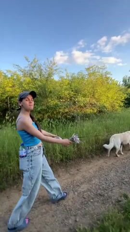 The video shows a young woman walking a white dog on a dirt path. The woman is wearing a blue tank top, loose-fitting light blue jeans, and a dark baseball cap. She's holding the dog's leash in her right hand, and there appears to be a water bottle in her back pocket. The dog is walking to her right, staying on the path. The background consists of green trees and grass, and the sky is blue with scattered clouds.