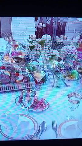 The video shows a long table set for a meal. The table is covered with a white tablecloth and adorned with white flowers. On the table are plates with food, glasses, and cutlery. In the foreground, there are plates with folded white napkins and silverware. A glass pitcher filled with a red liquid is on the left side of the table. A platter of sliced meats and garnishes is in the center of the table. There are also smaller dishes of food and drinks. In the background, there are chairs and decorative elements. A hand reaches in to pour liquid from the pitcher. The video captures the detailed setting of a formal meal, emphasizing the presentation of the food and the elegant table arrangement.