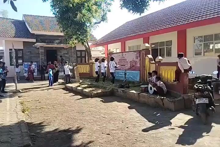 A group of children in school uniforms are gathered outside a building. Some children are dancing in a line, while others are seated on a low wall, playing musical instruments. A banner hangs behind the dancing children. There are other people standing around, observing. A motorcycle is parked to the right of the frame. The ground is covered in dirt and gravel, and there is some vegetation visible. The building appears to be a school, with a red roof and white walls. The sky is clear and sunny.