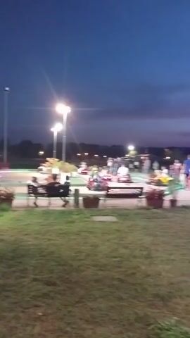 The video shows a nighttime scene at what appears to be an amusement park or fair. There are various attractions visible, including what looks like a carousel with lights and a ride with flashing lights in the background. People are scattered throughout the area, some walking, some sitting. The lighting is a mix of artificial lights from the attractions and streetlights, illuminating the dusk sky. The foreground is a grassy area with some potted plants and benches. The camera perspective is low, looking towards the attractions.