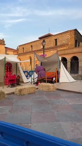 The video shows an outdoor scene with several people interacting around makeshift stalls. In the foreground, a person in a purple tunic and red leggings is shown leaning over a structure with a tan canvas roof, appearing to adjust something. To the left, hay bales are visible, along with a white tent. In the background, a historical-looking brick building stands, with arched openings on the ground floor. Several other people are positioned around tables and display areas, some wearing period costumes in shades of white and blue. One person is seen interacting with someone at a table covered with items. The ground is paved with large square stones. The overall impression is a historical reenactment or festival setting.