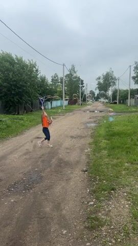 A young girl in an orange shirt and dark shorts stands on a dirt road and blows bubbles with a wand. She is facing away from the camera, looking down the road. The road is lined with grass and trees, and there are houses and fences on either side. The sky is cloudy. Bubbles are scattered throughout the frame.