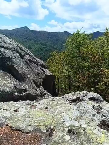 The video shows a view from a rocky landscape toward rolling mountains under a blue sky with scattered clouds. The foreground features large, gray rocks covered with patches of lichen. Behind the rocks, the landscape opens to reveal a valley and a series of green, forested mountains receding into the distance. The sky is bright blue with puffy white clouds adding depth to the scene. A few trees are visible on the right side of the frame, partially obscuring the view. The video is taken from a stationary position, providing a panoramic view of the natural landscape.