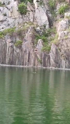 A person in a dark swimsuit dives into a murky green lake from a rocky cliff. They create a splash and then swim away from the cliff as another person wearing a dark swim cap swims nearby. The cliff is steep and has a ladder attached to it. It also has some green foliage growing on it.