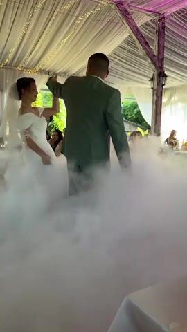 The video captures a wedding ceremony taking place outdoors under a canopy decorated with white drapery and fairy lights. A bride in a white off-the-shoulder wedding dress and a groom in a green suit are dancing. They are surrounded by a dense cloud of fog, creating a dramatic visual effect. Guests are seated at tables in the background, some of whom are looking on and taking photos. The scene appears to be set during daylight hours. The wedding is likely taking place in Arbanassi, Bulgaria, as indicated by the location context. The mood seems celebratory and romantic, with the couple appearing to be enjoying their first dance. No visible text is present in the video.