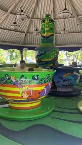 The video shows a Scooby Doo-themed teacup ride spinning at a park. The teacups feature different characters and scenes from the cartoon, such as Scooby Doo, Shaggy, and haunted castles. Several people are riding the teacups, including children and adults. The ride is covered by a white canopy, with light fixtures hanging from the ceiling. The platform and teacup bases are painted in green and yellow colors.