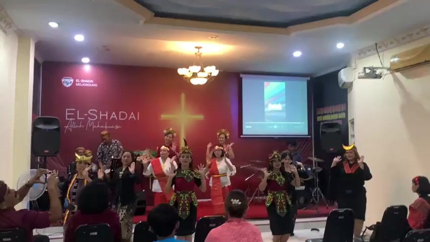 A group of women are dancing on a stage in a room. They are wearing traditional Indonesian clothing. There is a cross on the wall behind them. There is also a projector screen showing an image. There are people sitting in chairs watching the performance.