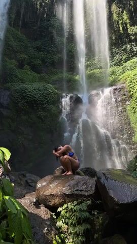 A woman in a blue bikini squats on a rock in front of a multi-tiered waterfall. The waterfall is surrounded by lush green foliage. The woman is facing away from the camera and her hands are in her hair. The lighting suggests it is daytime and sunny.