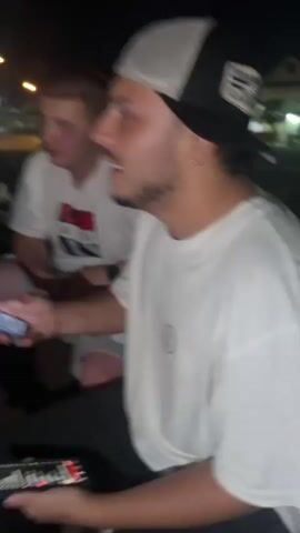 Two young men are sitting outdoors at night. The man in the foreground is wearing a black and white trucker hat, a white t-shirt with a smiley face logo, and dark pants. He is holding a smartphone in his left hand and gesturing with his right hand while speaking. The man in the background is wearing a white t-shirt with a graphic design. He is holding a smartphone in his left hand and looking towards the man in the foreground. The environment appears to be a street or parking lot with some buildings and streetlights visible in the background. The lighting suggests it is nighttime.