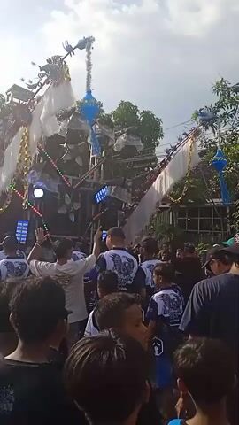 A crowd of people are enjoying a loud music event, likely a parade or mobile music gathering. They are assembled around a large, decorated vehicle, possibly a truck or float, adorned with lights and white flags or banners. The music is accompanied by flashing lights, creating a dynamic visual display. Many attendees are wearing similar shirts, suggesting they are part of a group or organization. The setting appears to be a residential area, with trees and houses visible in the background. The overall atmosphere is festive and energetic.
