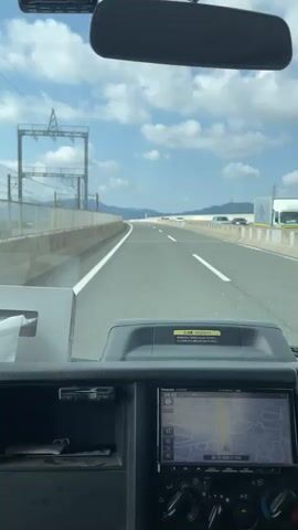 The video provides a first-person perspective from inside a vehicle, likely a truck, as it drives on a highway. The view is dominated by the windshield, revealing a clear blue sky with scattered clouds. The highway stretches ahead, with a white line marking the lanes. On the left side of the road, there is a guardrail and overhead electrical infrastructure, possibly for a train line. In the distance, mountains can be seen.

The dashboard of the vehicle is visible in the lower part of the frame. A navigation system screen displays a map. There is also a rearview mirror mounted above the windshield, reflecting the sky and the interior of the vehicle. A tissue box is placed on the dashboard. The overall impression is of a smooth journey on a well-maintained highway on a sunny day.