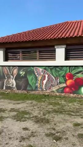 The camera pans across a mural on the side of a building. The mural includes images of a rabbit, a butterfly, a branch of cherries, a woodpecker, and a hedgehog. The building has a red tile roof and brown wooden trim. The ground in front of the building is dirt and grass. The sky is blue.