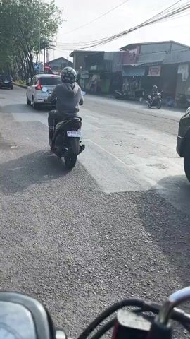 The video shows a street scene from the perspective of a rider on a motorcycle, looking forward. In the foreground, the handlebars of the motorcycle are visible. In the middle ground, a person on a scooter is riding in the same direction as the camera. Several cars are also on the road, moving in various directions. The environment appears to be a city street with buildings and trees lining the sides of the road. The sky is overcast. The road surface is asphalt and shows signs of wear and tear. Traffic lights are visible in the distance.
